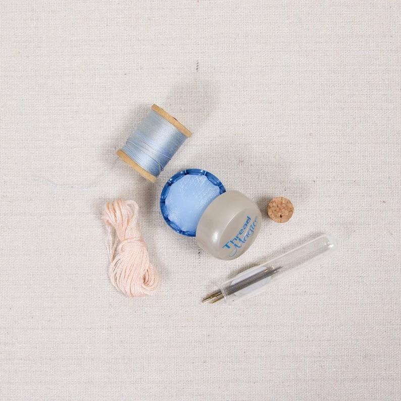 May include: An assortment of sewing supplies, including a spool of light blue thread, a bundle of pink thread, a blue wax container, a cork, and a needle tool. The items are arranged on a light-colored fabric background.