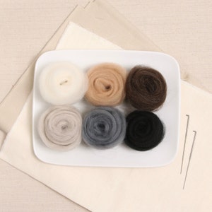 May include: Six balls of wool yarn in various colors, including white, beige, brown, gray, and black, arranged on a white plate. Two felting needles are on a beige fabric background.