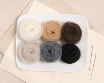 May include: Six balls of wool yarn in various colors, including white, beige, brown, gray, and black, arranged on a white plate. Two felting needles are on a beige fabric background.