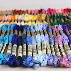 May include: A collection of embroidery floss spools in various shades of blue, from dark navy to light sky blue. The spools are arranged in a rainbow pattern, with the darkest shades at the bottom and the lightest shades at the top. The spools are labeled with numbers, such as 806, 820, 823, 750, 798, 932, 162, 157, 150, 141, and 155.