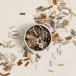 May include: A close-up of a glass jar filled with small, gold, brown, and silver glass beads. The jar is sitting on a beige fabric surface. The beads are scattered around the jar.