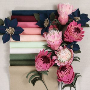 May include: A collection of felt fabric swatches in various shades of brown, pink, and blue, with a bouquet of pink felt protea flowers arranged on top.