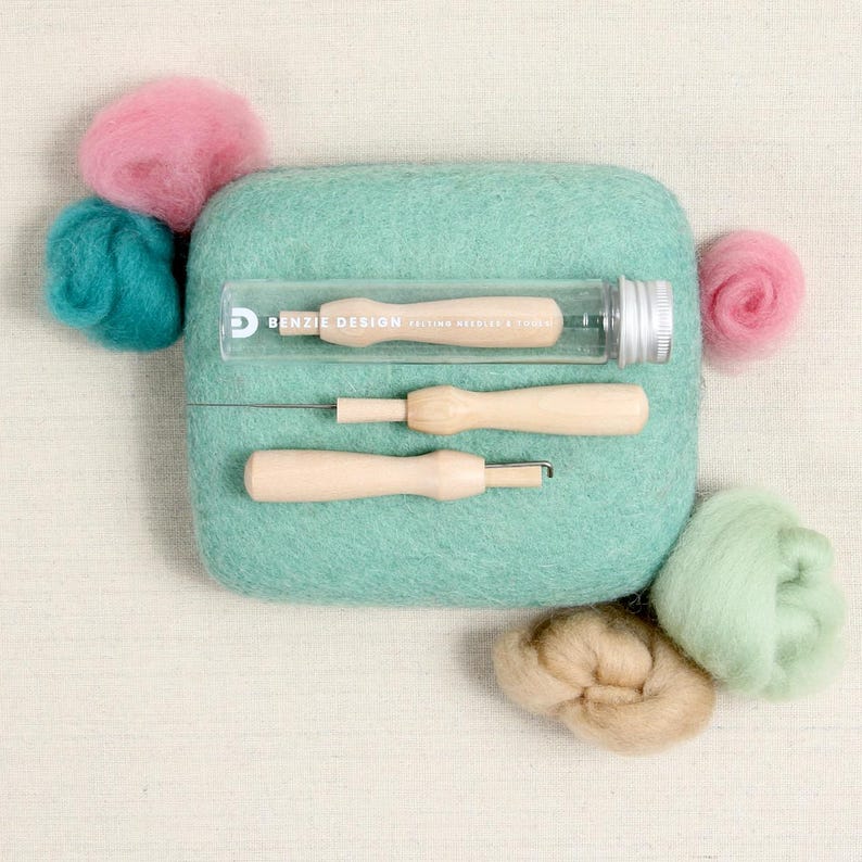 May include: A felting kit with two felting needles, a tube of felting needles, and five balls of wool in various colors: pink, teal, light green, beige, and light pink. The kit is on a light beige fabric background.