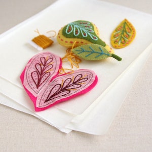 K&ouml;nnte beinhalten: Ein Set Filz-Ornamente mit einem rosa Herz mit braunen Stickereien, einer gelben Birne mit gr&uuml;nen Bl&auml;ttern und einem gelben Blatt mit gr&uuml;nen Stickereien.
