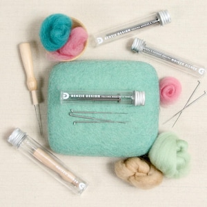 May include: A set of felting needles and tools in clear tubes with a green felting pad, pink, green, and beige wool roving, and a wooden felting needle. The tubes are labeled "Benzie Design Felting Needles & Tools".