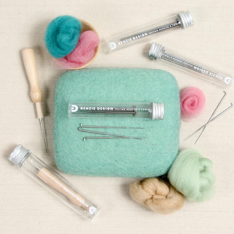 May include: A set of felting needles and tools in clear tubes with a green felting pad, pink, green, and beige wool roving, and a wooden felting needle. The tubes are labeled "Benzie Design Felting Needles & Tools".