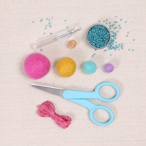 May include: A craft supplies set with a variety of colors. The set includes a small bowl of turquoise beads, a pair of blue scissors, a pink ball of yarn, four felt balls in pink, yellow, blue, and purple, and a glass tube with needles.