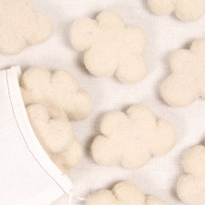 Może przedstawiać: Zestaw dziesięciu chmur z filcu w kolorze kremowym. Chmury są ułożone na jasnobeżowym tle z tkaniny.