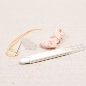 May include: A set of embroidery tools including a needle threader, a small spool of pink embroidery floss, a clear plastic needle minder, and a yellow thread.
