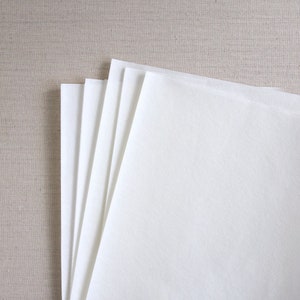 K&ouml;nnte beinhalten: Ein Stapel wei&szlig;er Papierb&ouml;gen, in der Mitte gefaltet, auf einer beigefarbenen, strukturierten Oberfl&auml;che.