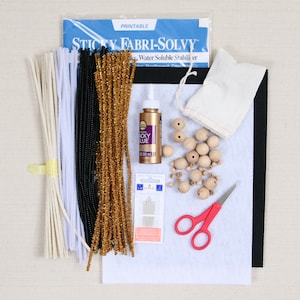 May include: A craft kit for making ornaments includes white, black, and gold pipe cleaners, wooden beads, a small white fabric bag, a bottle of tacky glue, a package of pins, a pair of scissors, and a piece of white felt.