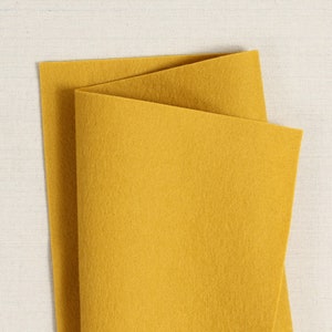 May include: A folded sheet of mustard yellow felt fabric. The felt has a soft, textured appearance and is displayed against a neutral, off-white background. The fabric is folded in a triangular shape, showcasing its thickness and colour.