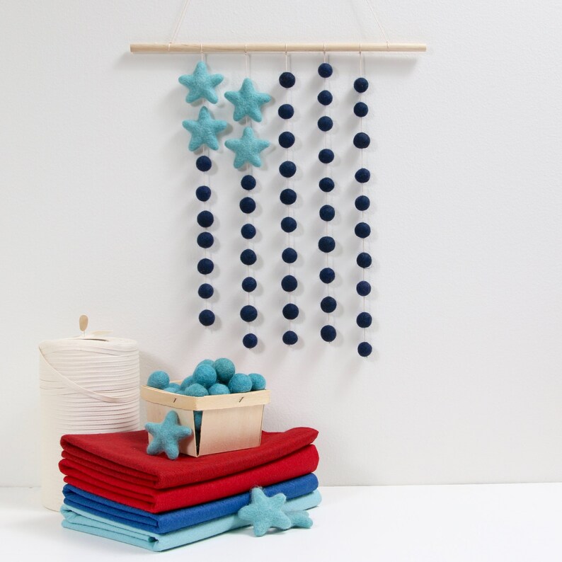 May include: A blue and white felt star garland with blue felt balls hanging from a wooden dowel. The garland is hanging on a white wall. There is a basket of blue felt balls and a stack of red, blue, and white fabric squares in the foreground.