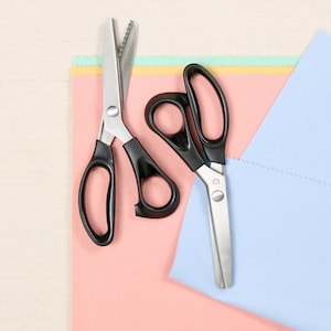 May include: Two pairs of scissors on a pink and blue fabric background. One pair is silver with a serrated blade and the other pair is black with a straight blade.