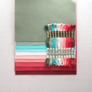 Puede incluir: Un conjunto de 12 carretes de hilo de bordar en tonos de verde, turquesa, blanco, rosa y rojo. Los carretes est&aacute;n dispuestos en una fila sobre una pila de cuadrados de tela de fieltro en tonos de verde, turquesa, blanco, rosa y rojo.