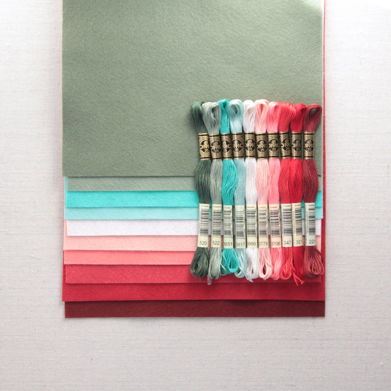 Puede incluir: Un conjunto de 12 carretes de hilo de bordar en tonos de verde, turquesa, blanco, rosa y rojo. Los carretes est&aacute;n dispuestos en una fila sobre una pila de cuadrados de tela de fieltro en tonos de verde, turquesa, blanco, rosa y rojo.