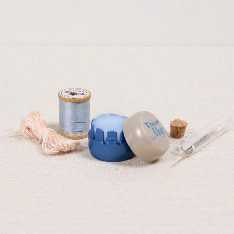 May include: A collection of sewing supplies, including a spool of light blue thread, a bundle of pink thread, a blue thread conditioner, a cork, and a needle case. The thread conditioner has the text "Thread Magic" on the lid.