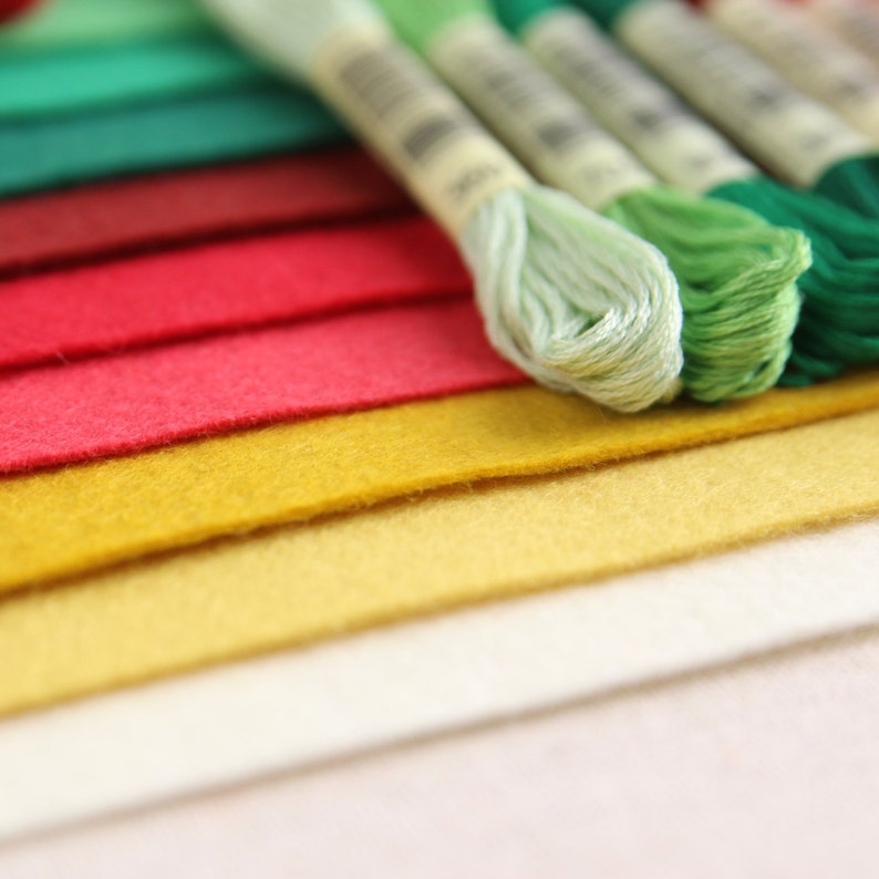 May include: A close-up of a stack of colourful felt fabric squares with green embroidery floss spools on top. The felt is in shades of green, red, yellow, and white.