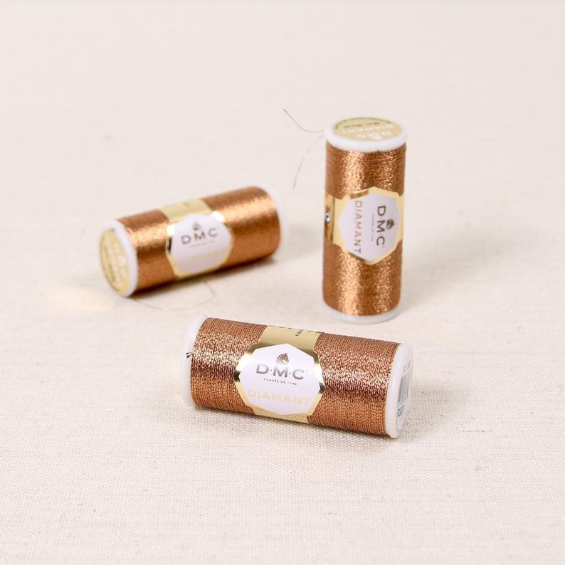 May include: Three spools of DMC Diamant embroidery thread in a metallic copper color. The spools are labeled with the DMC logo and the word "Diamant".