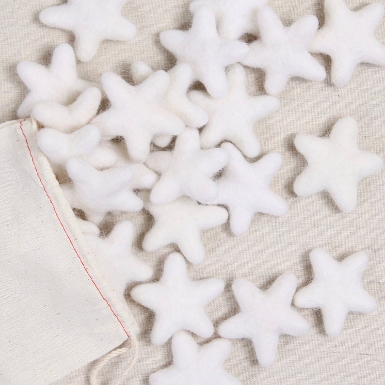 May include: A white canvas bag filled with white felt star ornaments. The stars are scattered on a beige fabric surface.