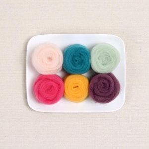 May include: A white rectangular plate holds six colorful wool roving balls. The balls are arranged in two rows of three, with colors including pink, teal, green, fuchsia, yellow, and purple. The wool appears soft and fluffy.