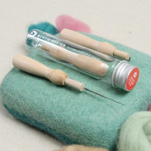 May include: A set of two felting needles in a clear glass tube with a silver lid. The needles have wooden handles and are used for needle felting. The tube has the text "Benzie Design Felting Needles & Tools" printed on it.