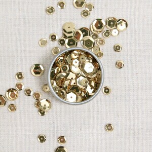 May include: A top-down view of a small, round, silver tin filled with gold sequins. The sequins are scattered around the tin on a beige, textured surface. The sequins are hexagonal in shape and have a shiny, metallic finish.