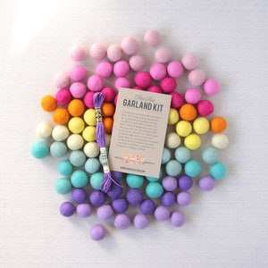 May include: A DIY felt ball garland kit with a rainbow color gradient. The kit includes a spool of purple embroidery floss and a card with instructions that reads "Felt Glam Garland Kit".