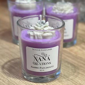 Peut inclure: Des bougies parfumées dans des verres transparents. La cire violette est surmontée de motifs floraux blancs. Chaque bougie porte une étiquette "Bougie Parfumée" et "Nana Creations Ambre Patchouli". Elles sont sur une surface en bois.