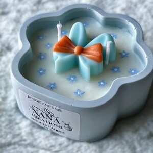 Peut inclure: Une bougie parfumée bleu clair en forme de fleur avec un nœud bleu clair sur le dessus. La bougie porte une étiquette blanche sur laquelle est inscrit "BOUGIE PARFUMEE NANA CREATIONS DOUCES PÂQUES".