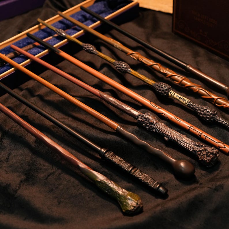 May include: A collection of wooden wands with intricate designs and varying colors, including brown, black, and green. Some wands have detailed handles and textured surfaces. The wands are arranged in a display case and on a dark surface.