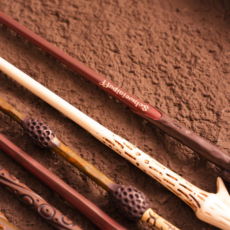 May include: A collection of wooden wands in various shades of brown and tan, with intricate carved details. One wand has the word "Schweineipuff" printed on it. The wands are arranged diagonally against a textured brown background.