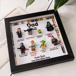 Op de afbeelding: Een ingelijst Star Wars-thema Lego-kunstwerk. Het zwarte frame omringt een witte achtergrond met Lego-figuren van personages als Darth Vader en Yoda. Tekst bevat "Dad" en "Yoda Best."