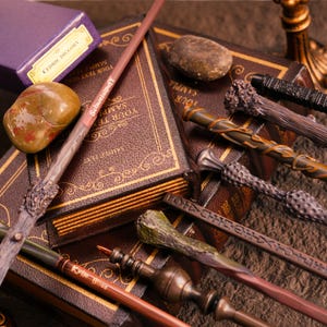 May include: A collection of detailed magic wands in various colors, including brown, green, and gold, are displayed on top of leather-bound books. A purple box labeled "Cedric Diggory" and a small stone are also present. Some wands have text that reads "Schweinipuff".
