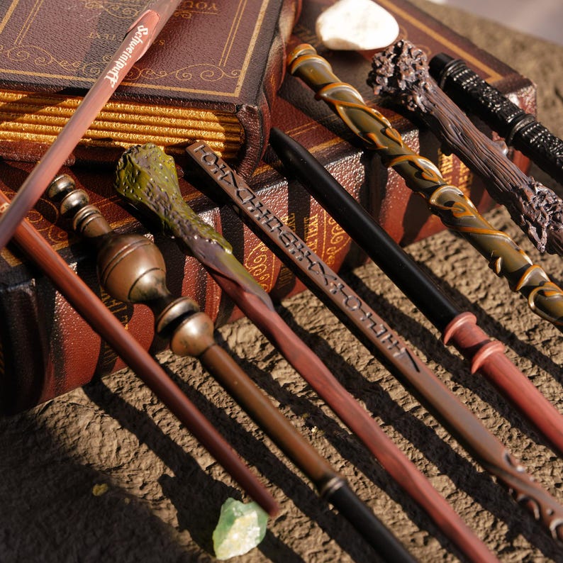 May include: A collection of intricately designed wands, each with unique details and colors, including brown, black, and green. Some wands have carved patterns and text. The wands are arranged near a leather-bound book.