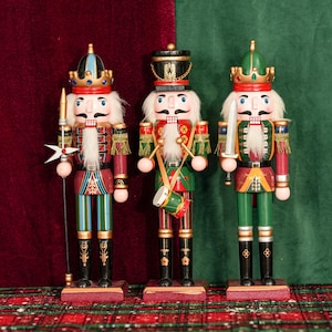 以下が含まれることがあります： 装飾的な木製のくるみ割り人形が3つ。各フィギュアは異なる制服を着ており、槍、ドラム、剣などのアクセサリーが付いています。くるみ割り人形は長方形の台座に立っており、伝統的なお祝いのテーマを体現しています。