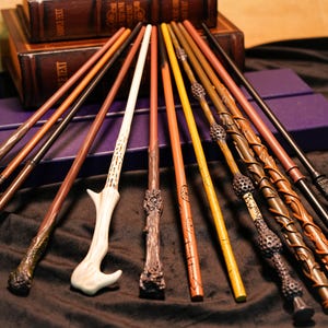 May include: A collection of various wizard wands in shades of brown, tan, and gold, with intricate designs and textures. Some wands have unique handles, and the wands are arranged on a dark surface near leather-bound books.