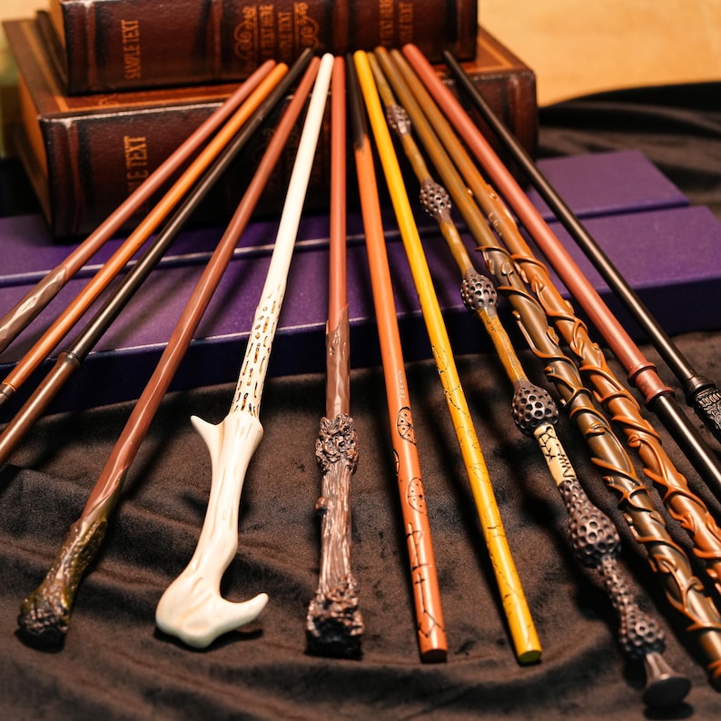 May include: A collection of various wizard wands in shades of brown, tan, and gold, with intricate designs and textures. Some wands have unique handles, and the wands are arranged on a dark surface near leather-bound books.
