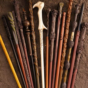 May include: A collection of various wands in shades of brown, black, and cream. The wands have unique designs, including carved details, swirling patterns, and textured grips. Some wands are straight, while others have curved or ornate handles.