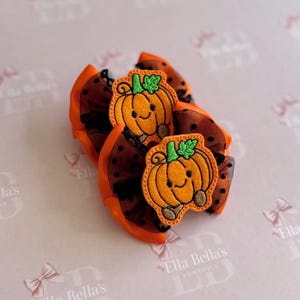 May include: A pair of hair bows featuring orange satin ribbon and black polka-dot tulle fabric. Each bow is centred with an embroidered felt patch of a smiling orange pumpkin with a green stem and leaf, and two small brown feet at the base.