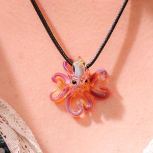 May include: A colorful glass octopus pendant necklace. The octopus is a mix of orange, pink, and purple hues, with black eyes and a clear head. The pendant hangs from a black cord necklace, creating a unique and eye-catching accessory.