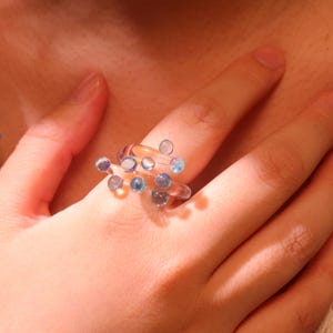 Può includere: Un anello trasparente e astratto con molteplici accenti sferici blu e trasparenti. L'anello è indossato su un dito, mostrando il suo design unico. Il design dell'anello è moderno e artistico.