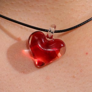 May include: A red heart-shaped glass pendant suspended from a black cord necklace. The pendant is transparent, allowing light to pass through, and is a vibrant shade of red. The necklace is worn around the neck.