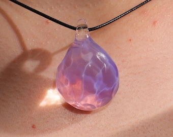 May include: A teardrop-shaped pendant necklace with a translucent, textured glass pendant. The pendant is a blend of pink and purple hues, suspended from a black cord. The glass has a unique, organic shape, and the overall design is simple and elegant.