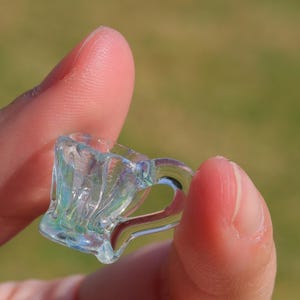 May include: A miniature, clear glass mug with a handle, held between fingers. The mug has a textured, rippled design and iridescent sheen, reflecting light with hints of blue and green. The mug is small, likely a decorative item.