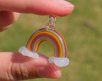 May include: A miniature glass rainbow pendant with a clear loop at the top and white cloud accents at the base. The rainbow features bands of red, orange, yellow, green, blue, and purple. The pendant is held between two fingers against a blurred green background.