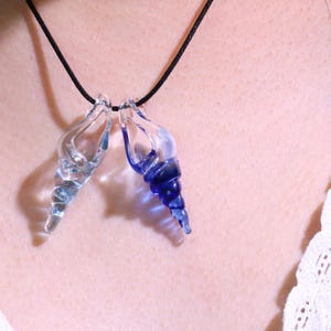 May include: Two glass seashell pendants, one clear and one blue, hang from a black cord necklace. The pendants are detailed with spiral designs and are worn against a person's skin.