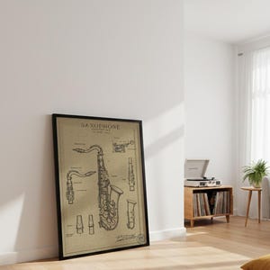 Puede incluir: Impresión enmarcada de estilo vintage de una patente de saxofón. La obra de arte presenta diagramas detallados y la palabra "SAXOPHONE" en la parte superior. La impresión está en un marco negro y se apoya en una pared blanca, mostrando un instrumento musical.
