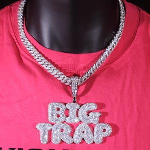 May include: A silver-coloured, iced-out chain necklace with a pendant reading "BIG TRAP" in large, bubble letters. The necklace is displayed on a red shirt.