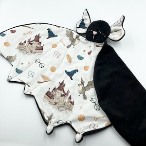 Könnte beinhalten: Eine schwarze, fledermausförmige Sicherheitsdecke mit einer weißen Stoffvorderseite, die ein Hogwarts-Schloss und andere Harry-Potter-Motive zeigt. Die Rückseite der Decke ist aus schwarzem Plüsch. Die Decke hat einen kleinen, plüschigen schwarzen Fledermauskopf mit rosa Ohren.